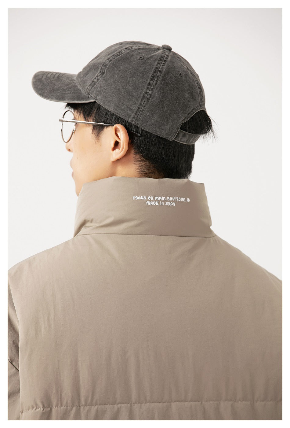 Rear view of a person wearing a taupe padded jacket with a high collar and a dark gray cap.