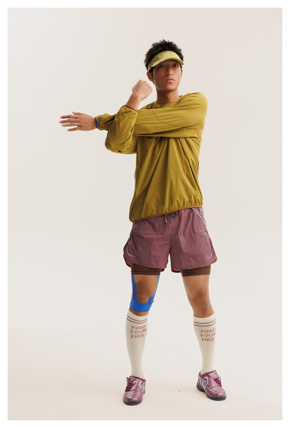 Model stretching in a mustard-yellow long-sleeve top, mauve running shorts, cream socks and a yellow visor.