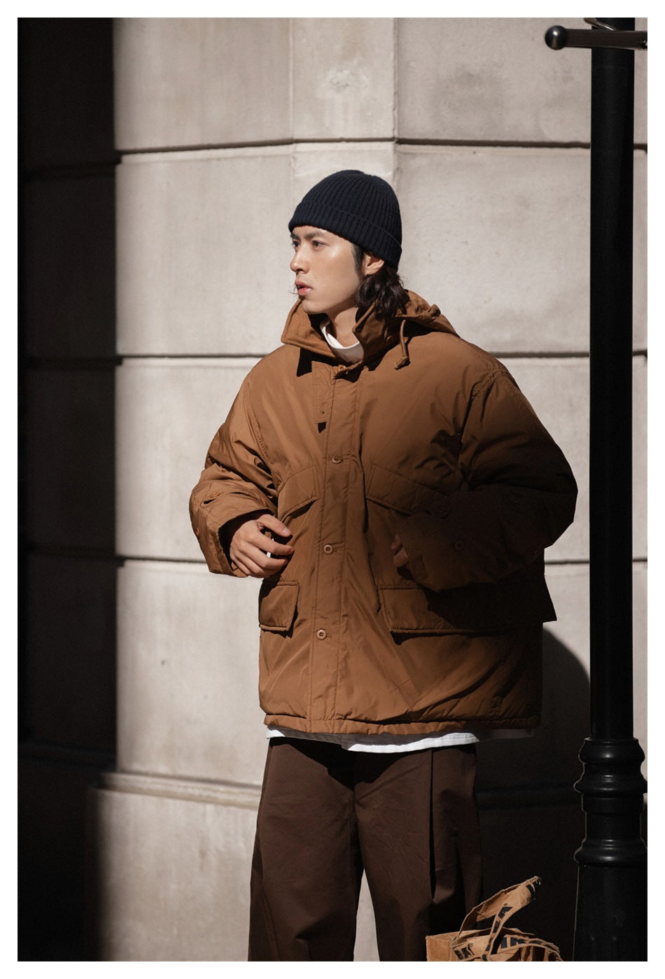 Model wearing the brown hooded jacket with large flap pockets, shown from a slight angle outdoors.