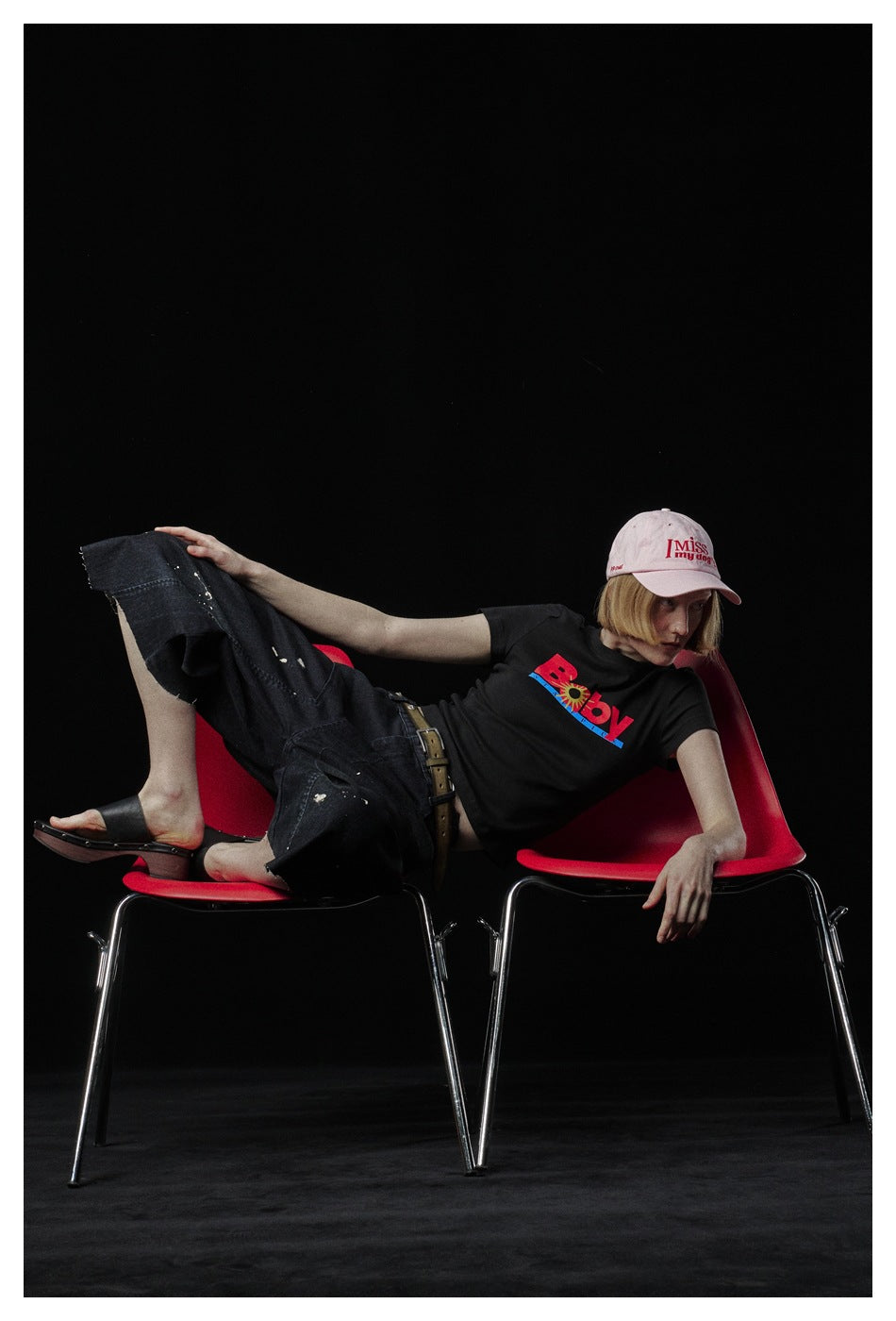 Model reclining across red chairs, wearing a black graphic T-shirt, distressed dark jeans, sandals, and a pink cap.