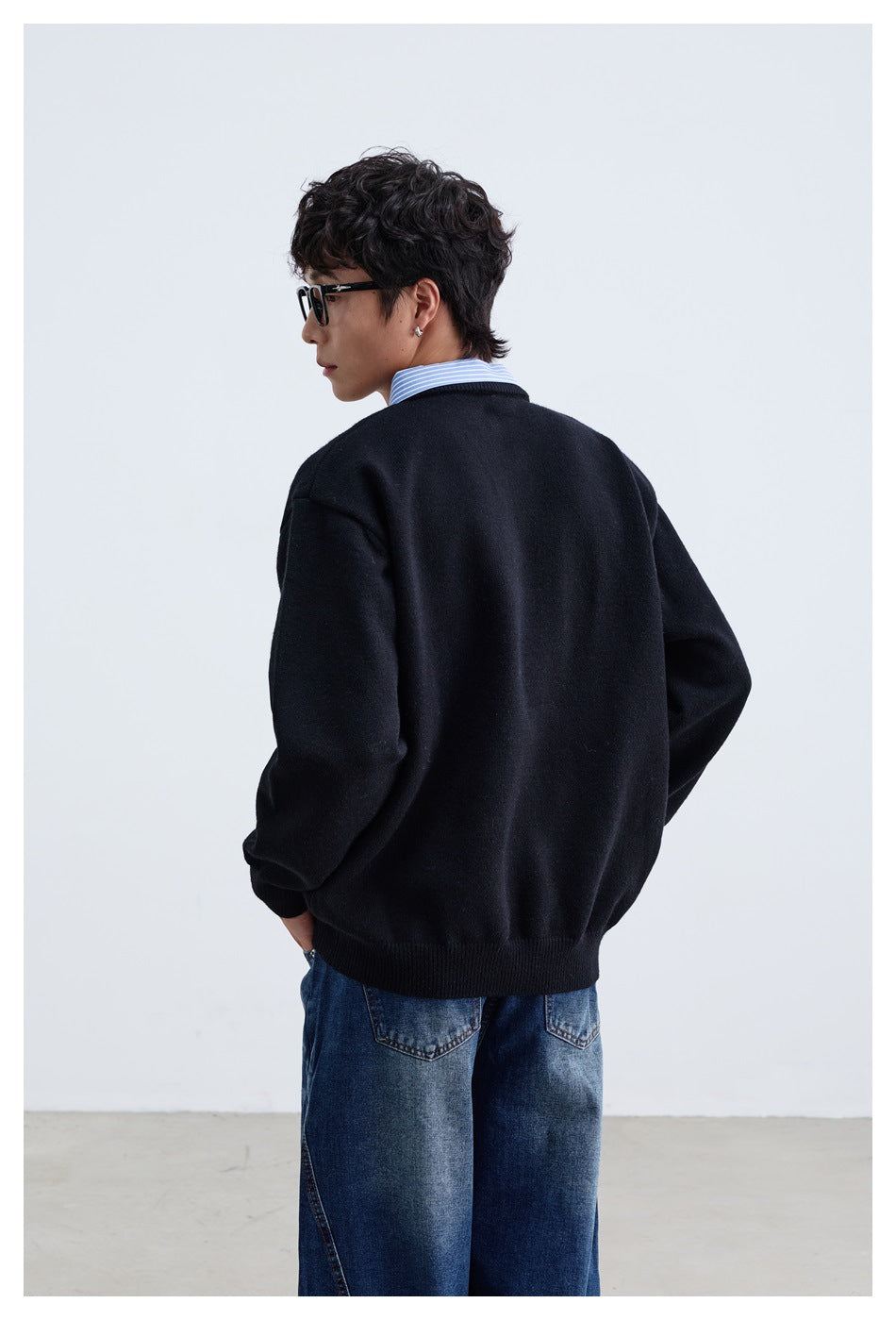 Rear view of a model wearing a black sweater with ribbed cuffs and hem, layered over a blue striped shirt.