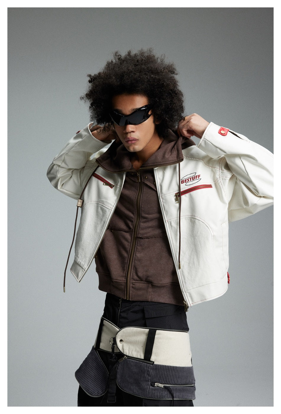 Model lifting the hood of a cream jacket with red zip pockets over a brown zip-up top.