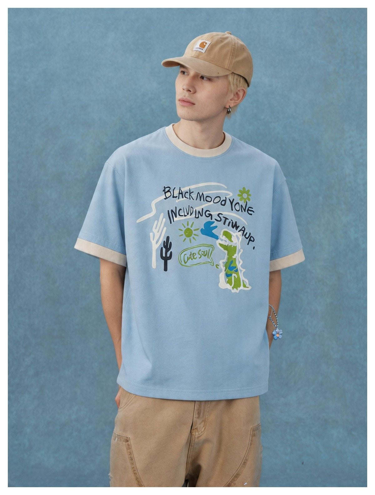 Model wearing a light blue ringer T-shirt with beige trim and graphic print, paired with tan pants and a cap