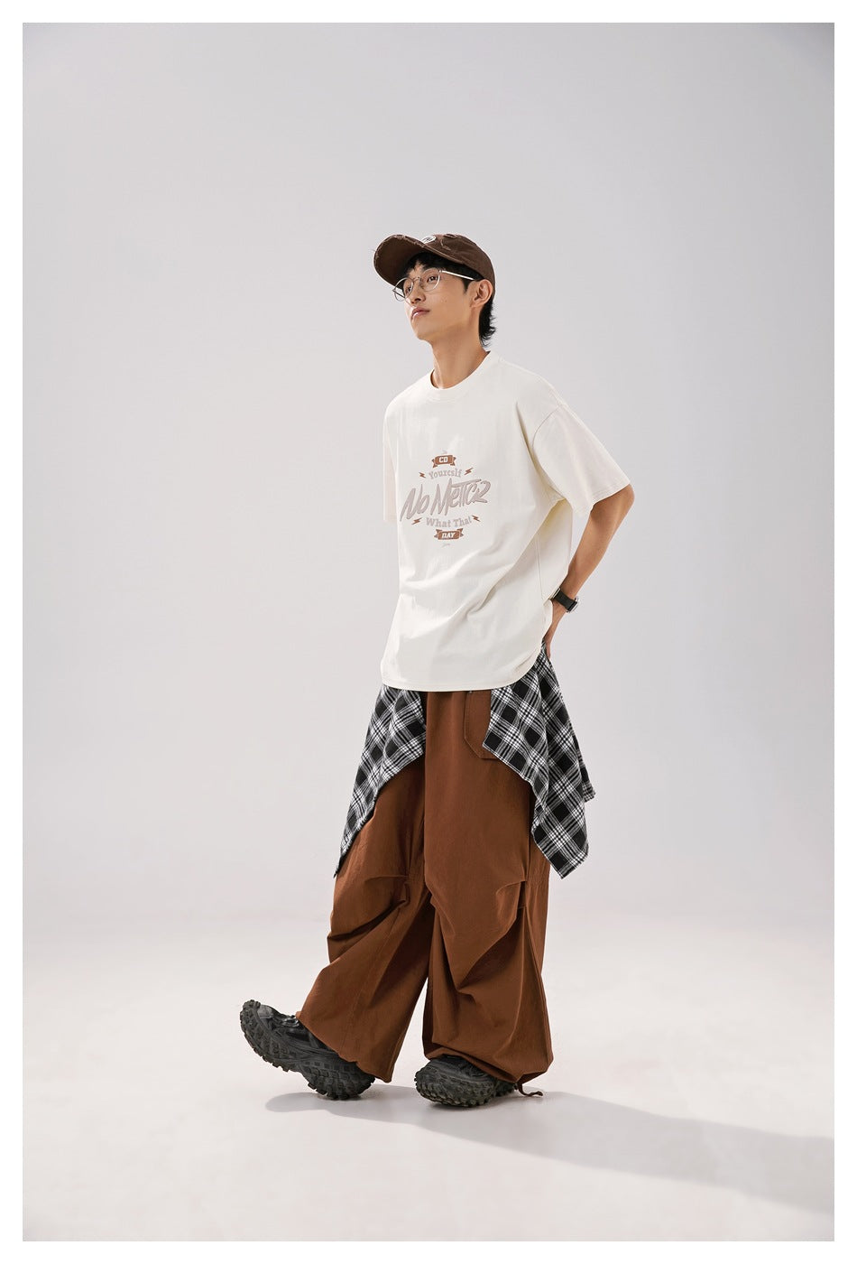 Model wearing an oversized ivory T-shirt with a brown and taupe chest graphic, styled with brown trousers and plaid fabric.