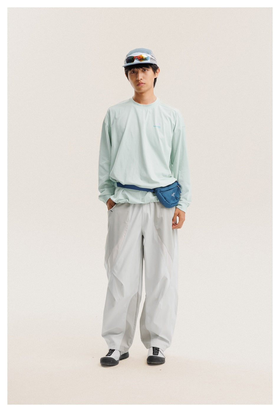 Front view of a man wearing a pale mint long-sleeve shirt and loose light-gray trousers, with a blue waist pouch.