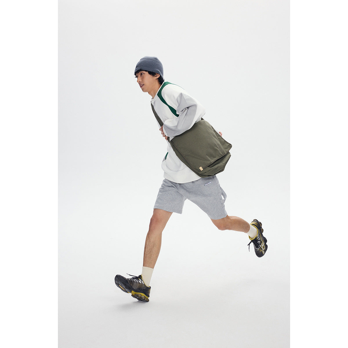 Model running in a white, green, and gray sweatshirt with gray shorts and a large olive shoulder bag.