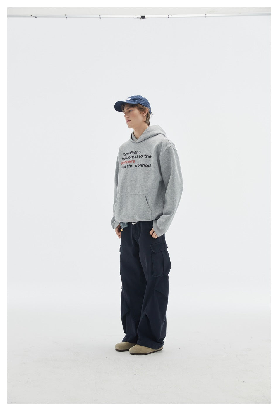 Three-quarter side view of a model in a gray printed hoodie, navy cargo pants, and blue cap.