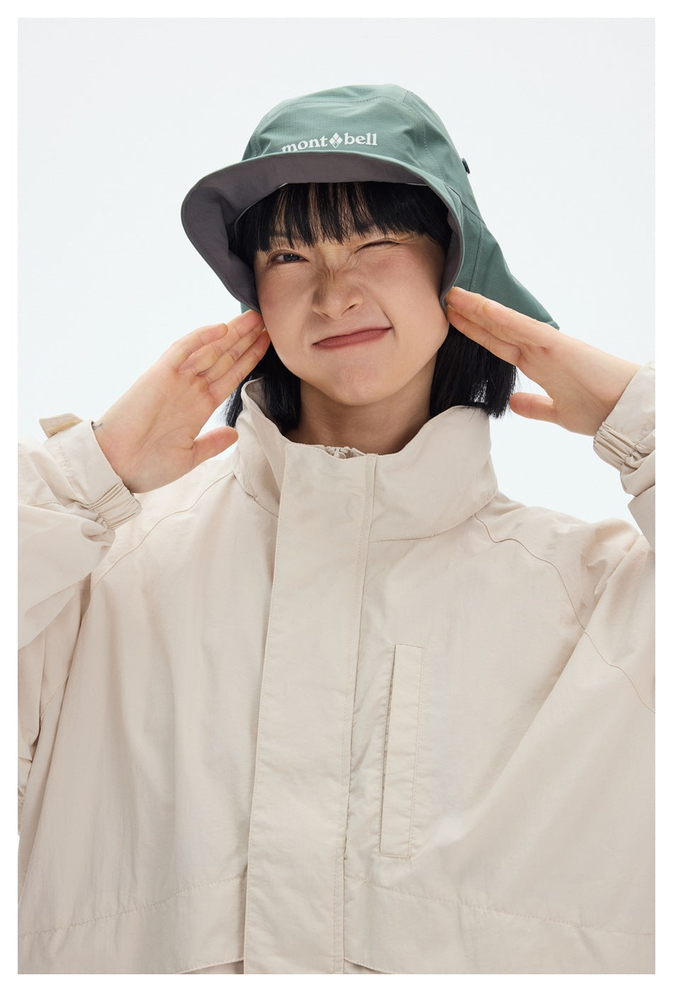 Close-up of a person in an off-white jacket with stand collar, chest pocket and elastic cuffs, wearing a green hat