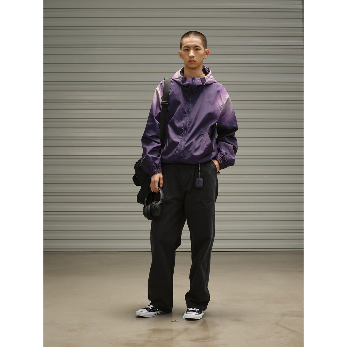 Man wearing a purple hooded zip jacket with elastic cuffs and loose black trousers, holding headphones.