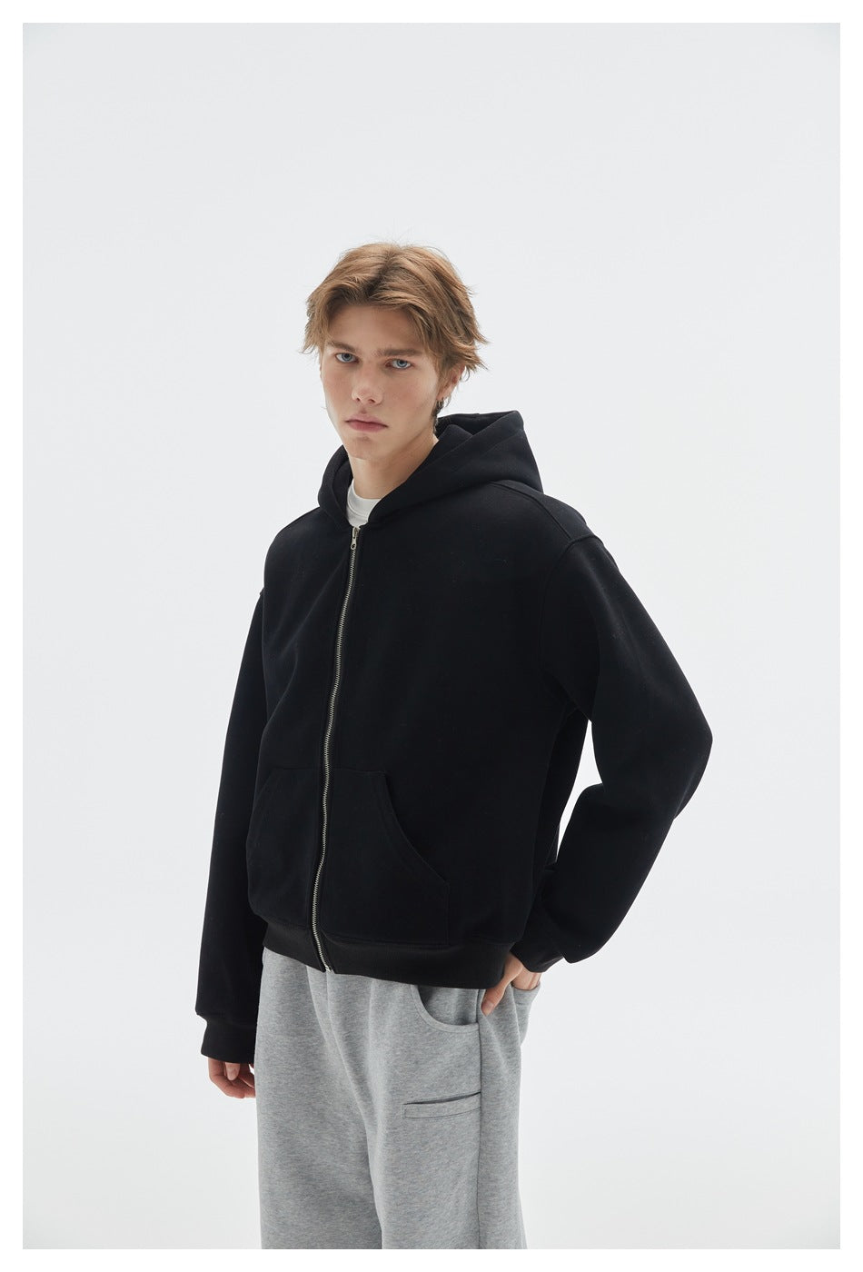 Model wearing a black zip-front hoodie with a hood, front pockets and ribbed cuffs, paired with gray pants.