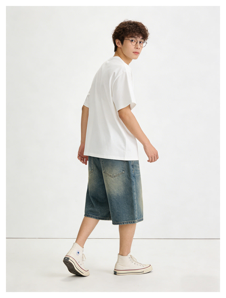Model turned to show the back of a plain white T-shirt and loose blue denim shorts.