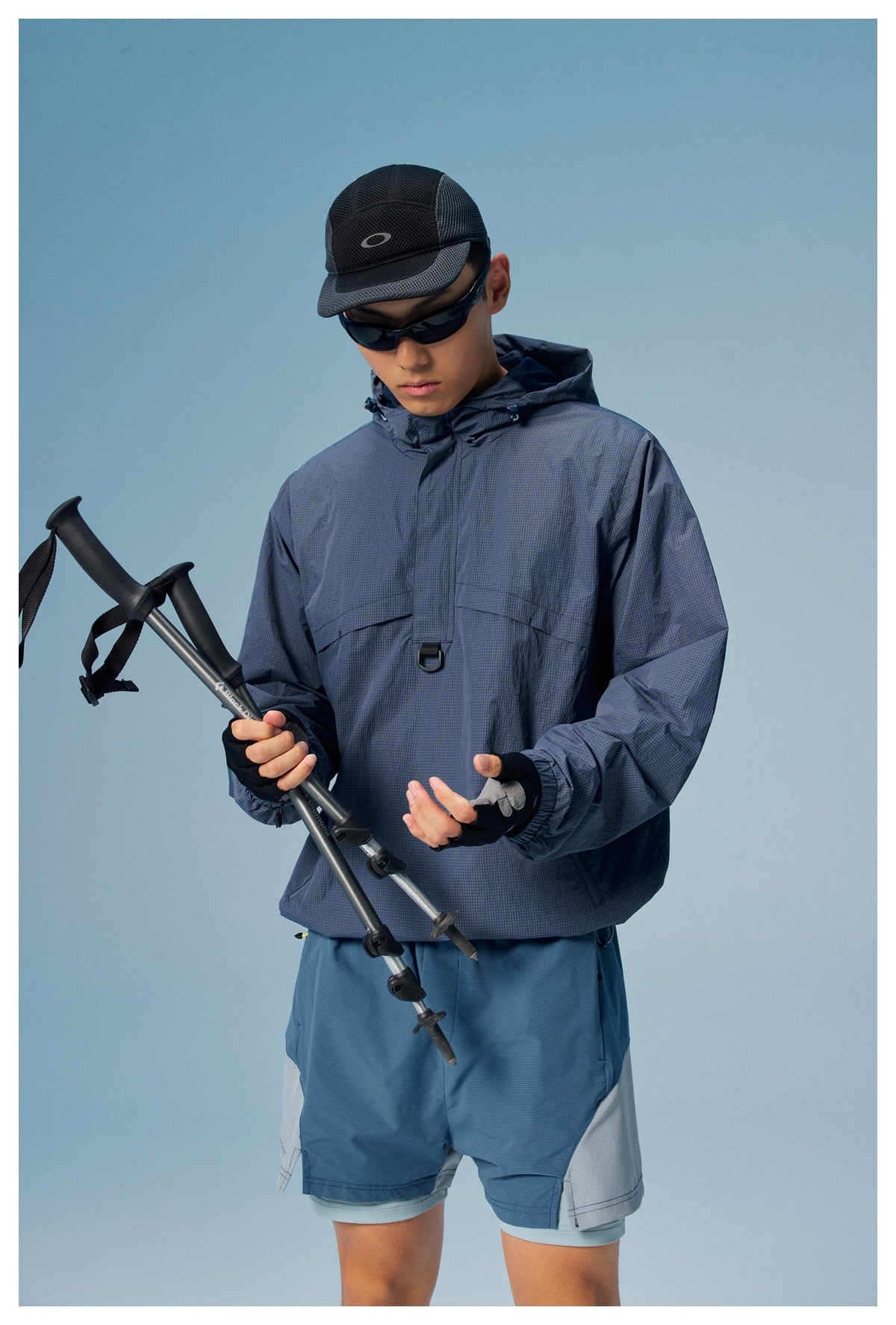 Model in a slate-blue hooded pullover jacket and blue-gray shorts holding trekking poles against a blue backdrop.