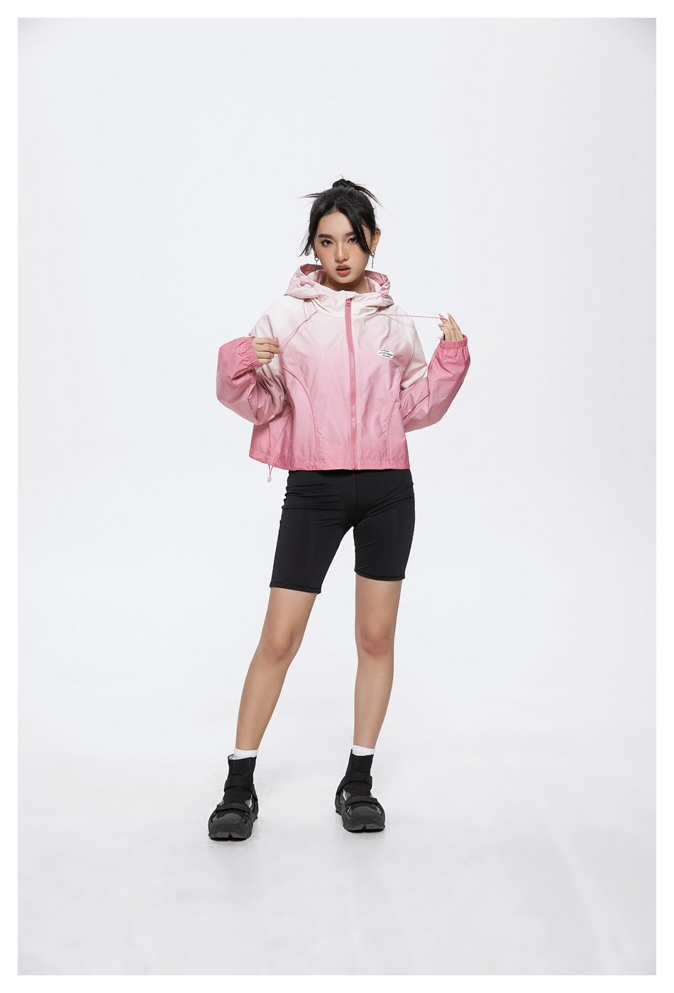Model wearing a pink gradient hooded zip jacket and black cycling shorts, holding the jacket's drawcords.