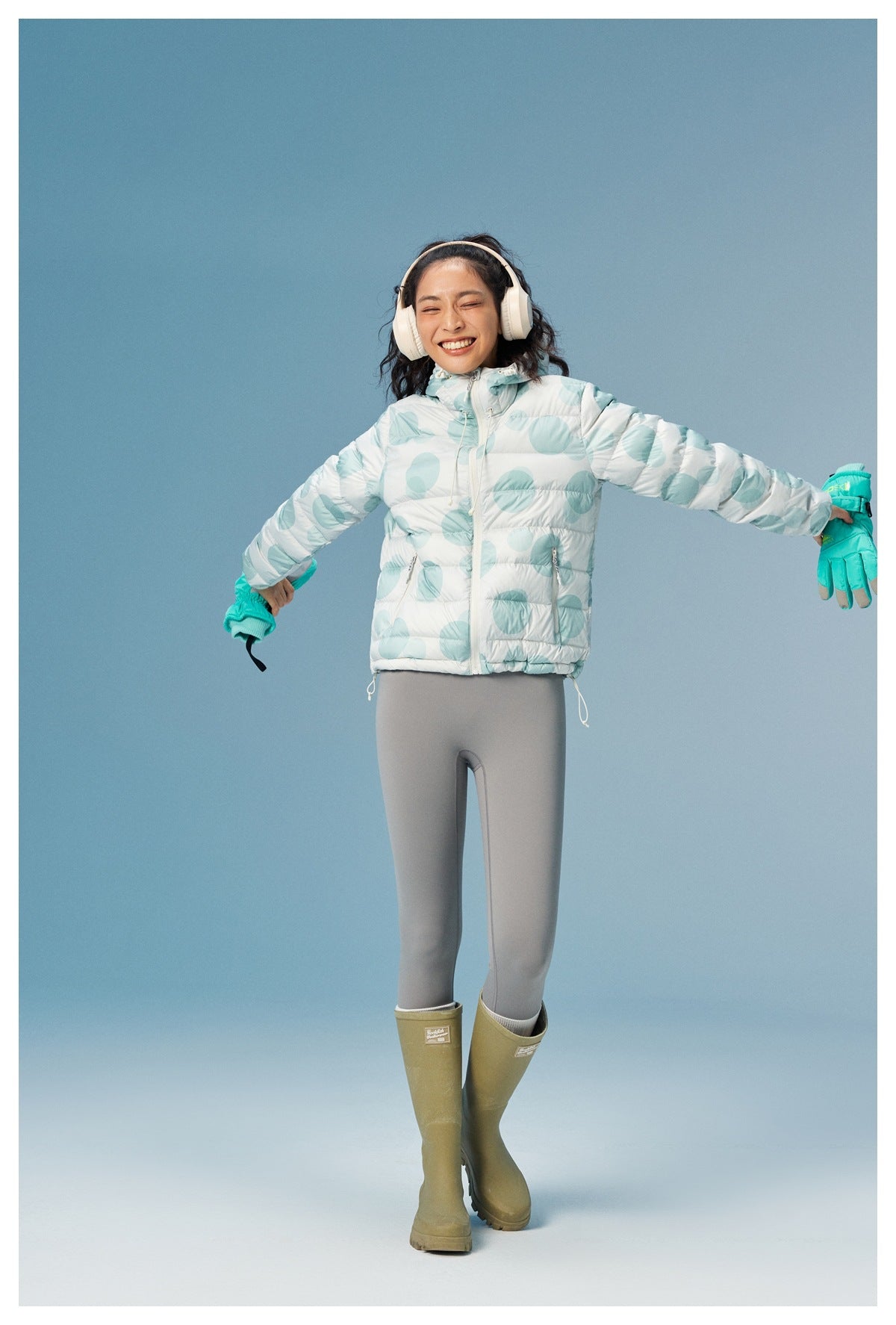 Front view of a model wearing a white quilted hooded jacket with aqua circular prints and gray leggings.
