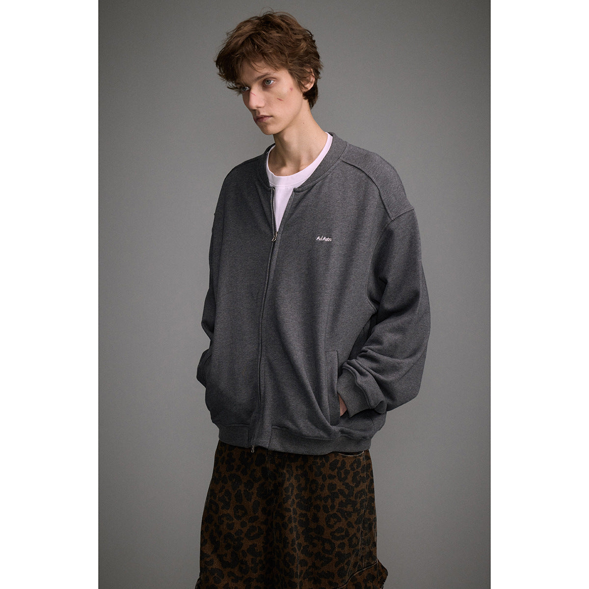 Model wearing a dark charcoal zip-front sweatshirt with a small chest logo and leopard-print trousers.