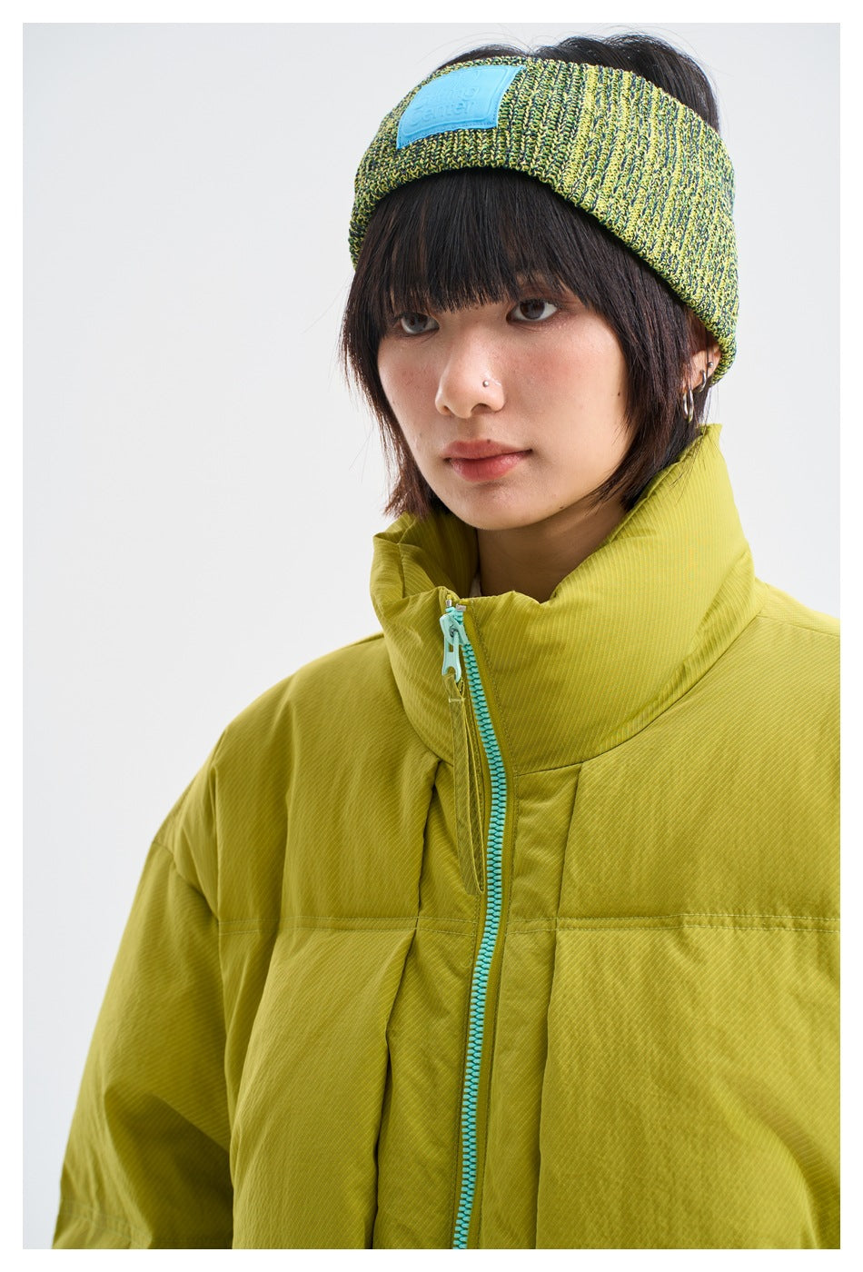 Close-up of a model wearing a chartreuse puffer jacket with a turquoise zipper and a green-yellow knit headband.