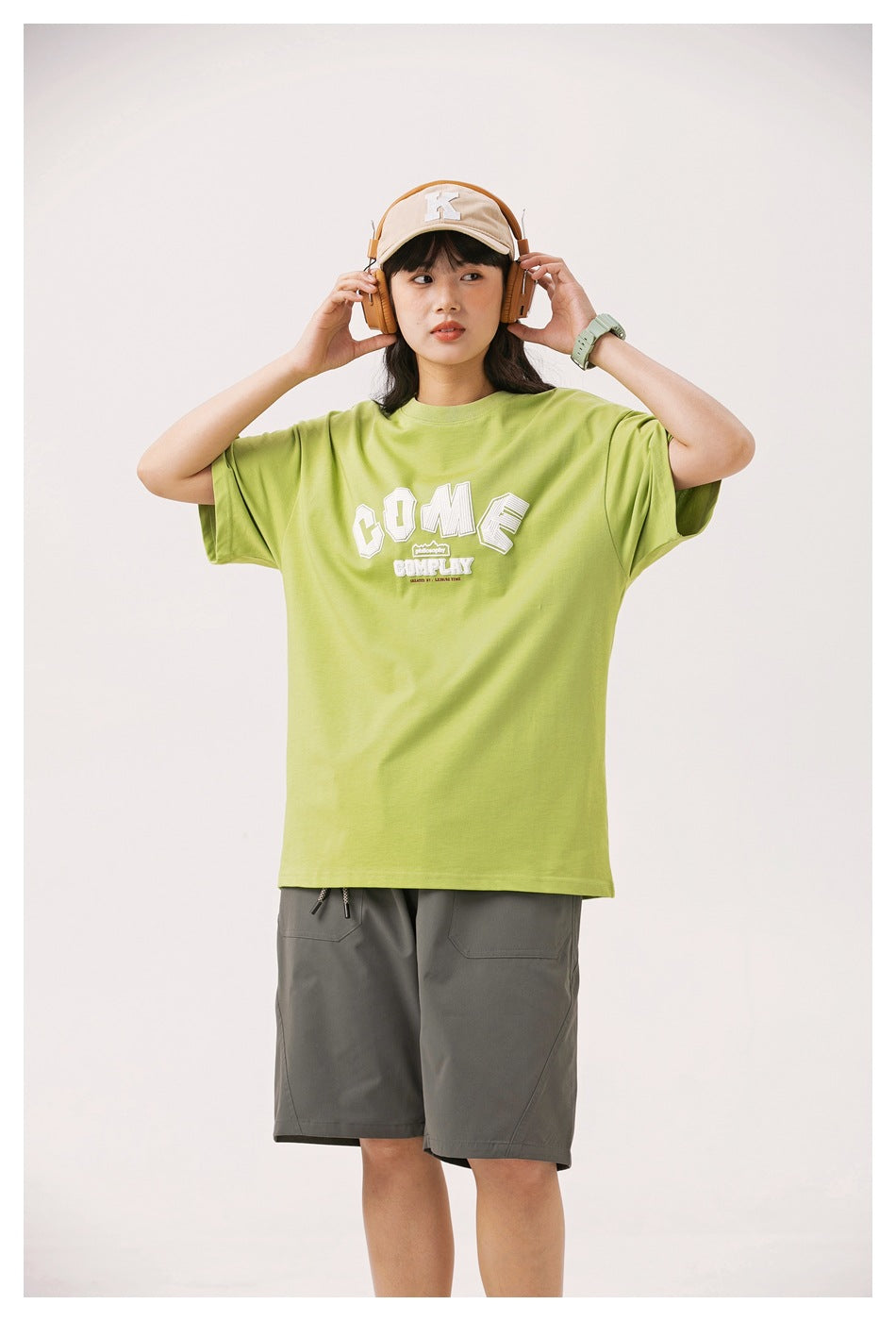 Front view of a model in a lime green oversized graphic T-shirt and gray shorts, adjusting her headphones.