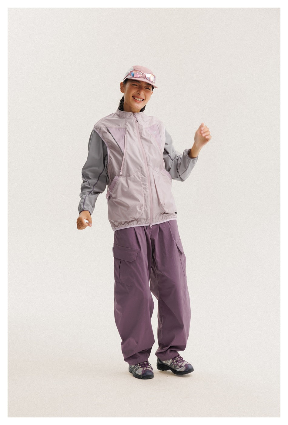 Angled view of a person wearing a pale mauve and gray utility jacket with plum cargo pants and a pink cap.
