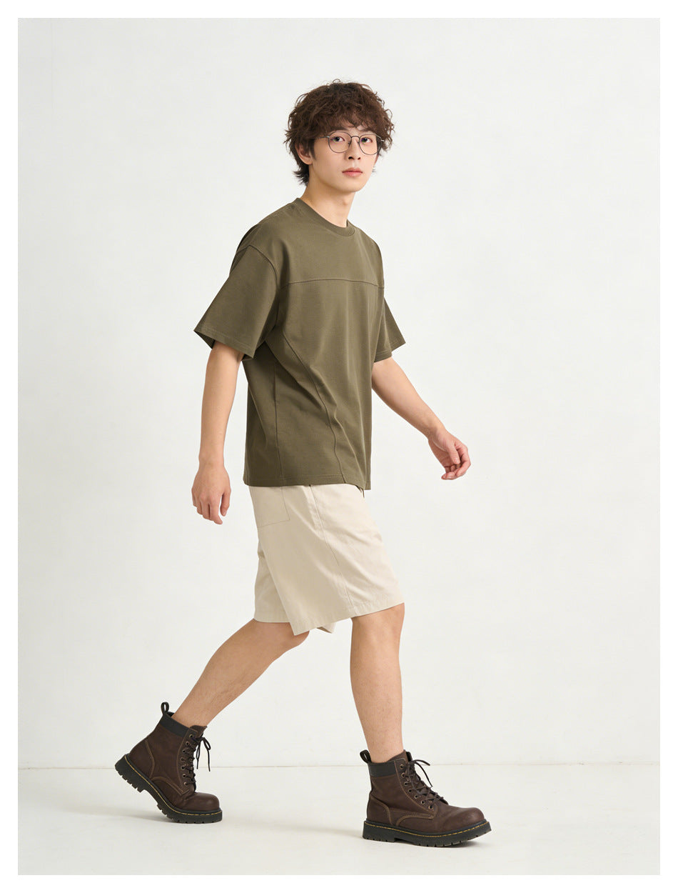 Model walking in an olive short-sleeve crew-neck T-shirt with a horizontal chest seam and beige shorts.