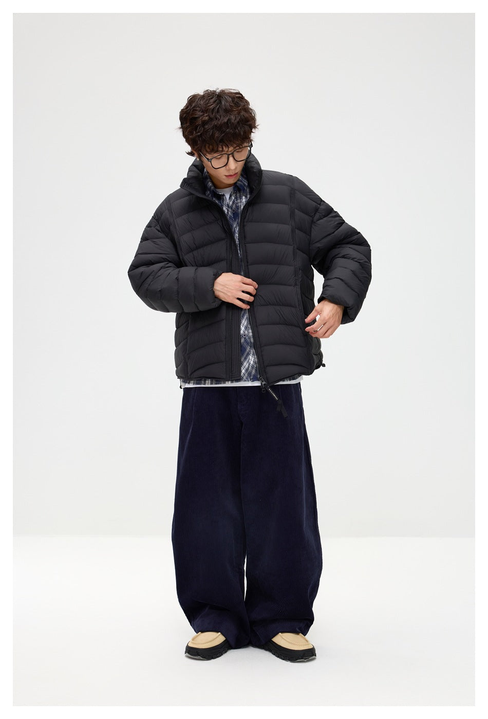 Model adjusting a black horizontally quilted puffer jacket with a raised collar, worn over a blue plaid shirt.