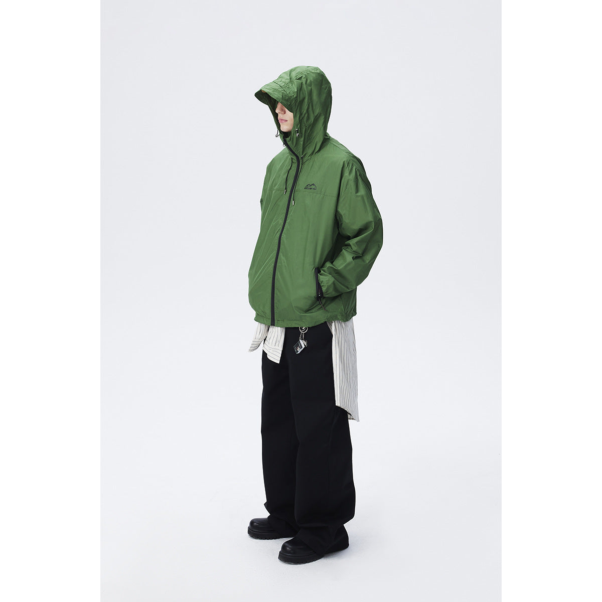 Model wearing a green hooded zip jacket with black trim, layered over a striped shirt and black trousers.
