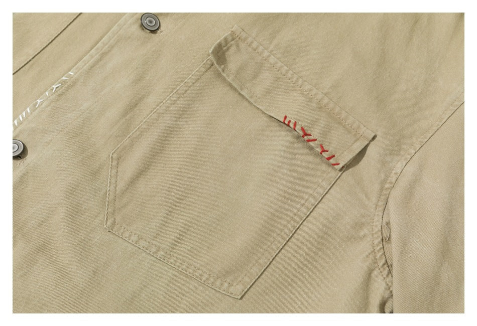 Close-up of a tan jacket with a buttoned flap pocket and contrasting red and white decorative stitching.