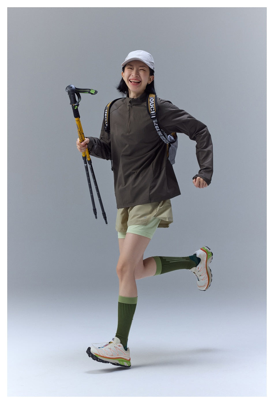 Model in a dark taupe quarter-zip top and layered shorts, smiling while holding hiking poles.