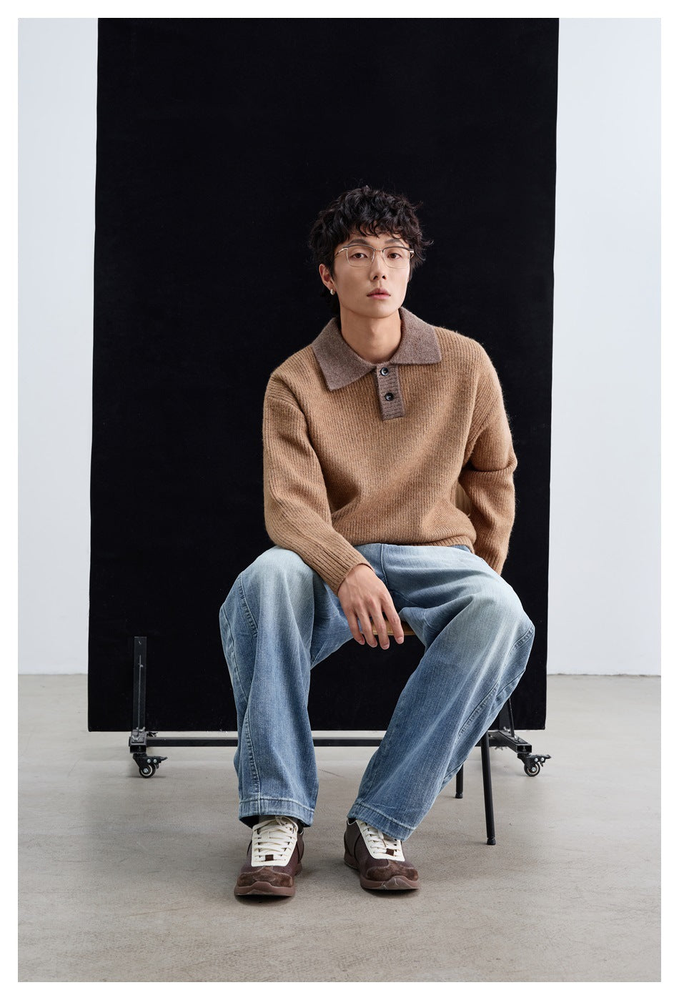 Seated model wearing a brown ribbed polo-collar sweater with light-wash wide-leg jeans and sneakers against a black backdrop