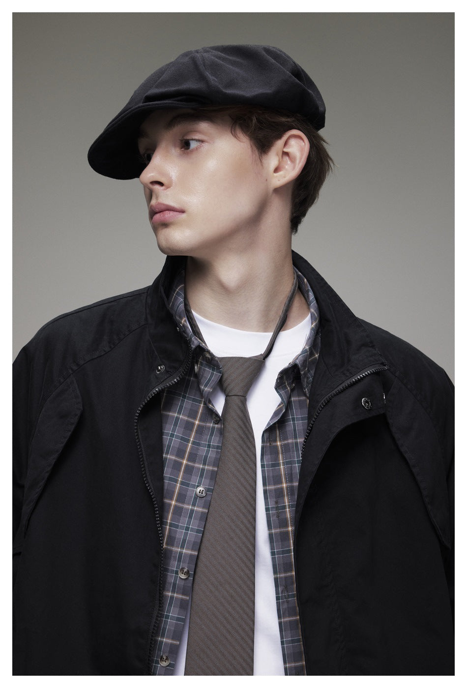 Close-up of a model in a black jacket and cap, layered over a plaid shirt, white tee and gray tie.