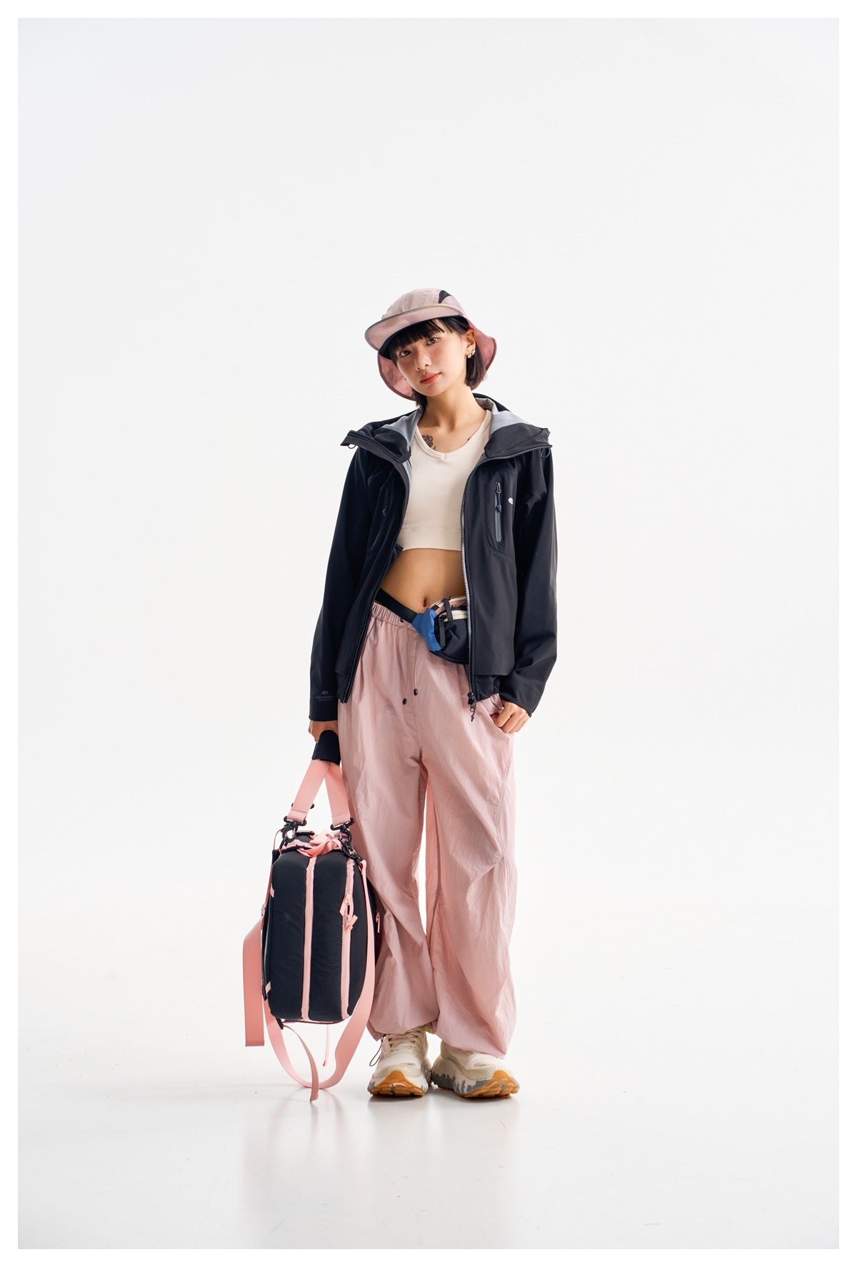 Model wearing a dark hooded jacket over a white crop top with loose pale pink pants, holding a black-and-pink bag.