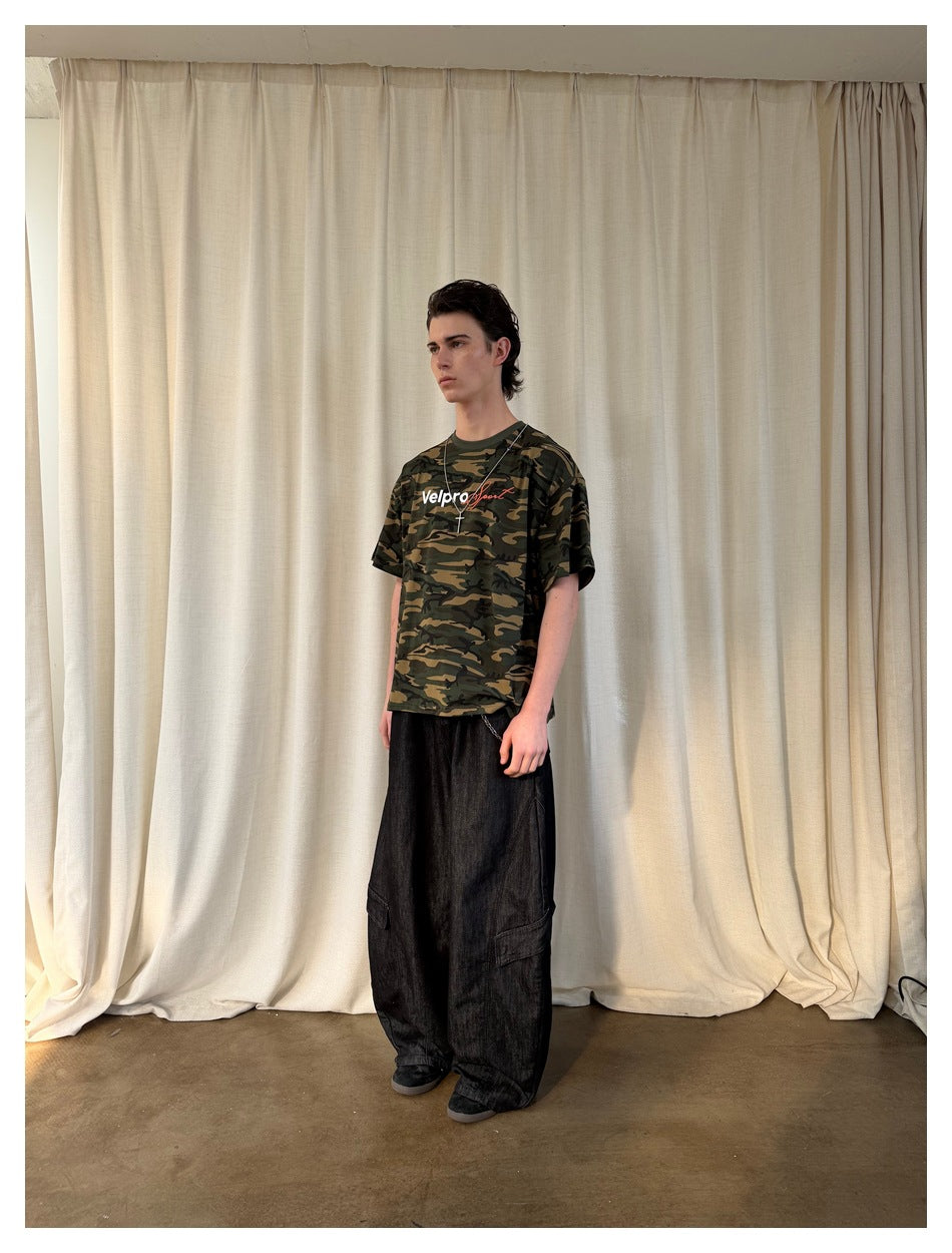 Three-quarter view of a model wearing a green and brown camouflage graphic T-shirt with loose black cargo jeans.