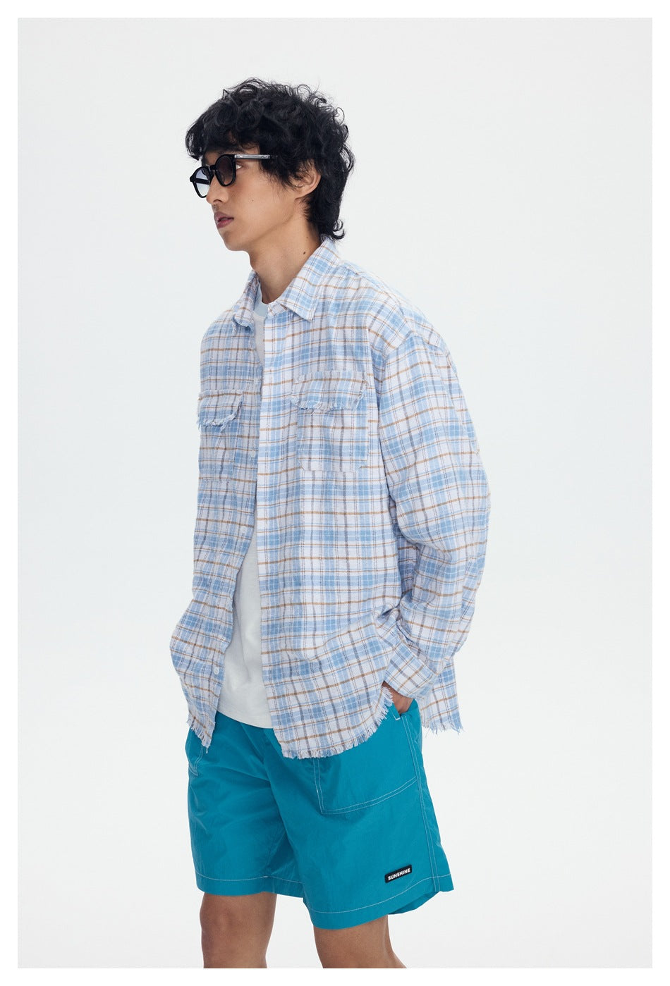 Three-quarter view of a model wearing a light blue plaid shirt with frayed edges and turquoise shorts.