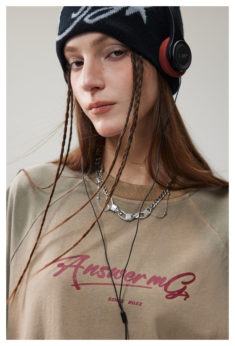 Close-up of an olive-gray T-shirt with burgundy script print, worn by a model with a black beanie and headphones.