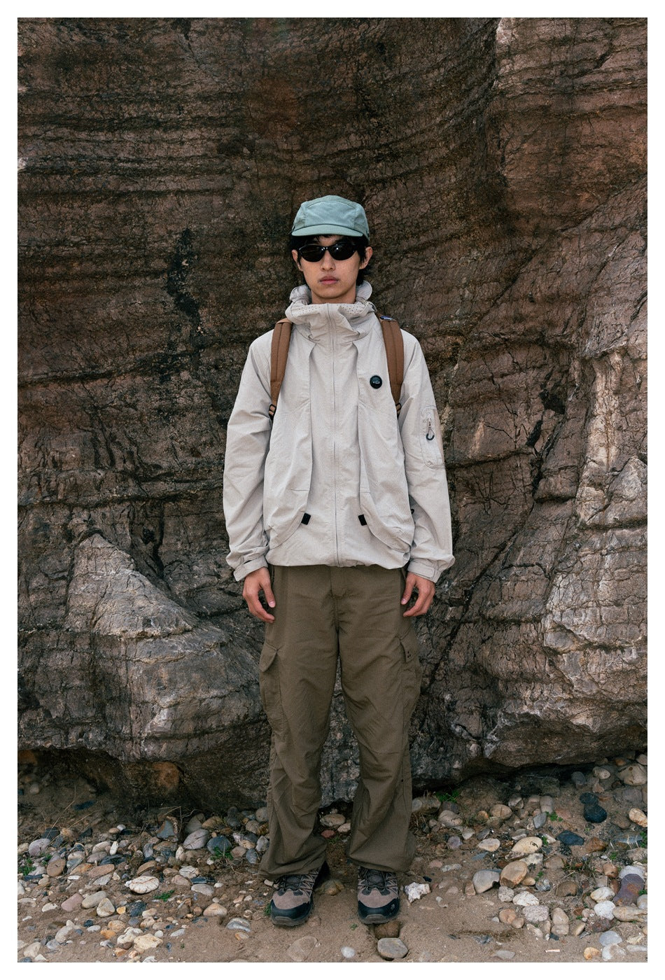 Person wearing a light beige hooded jacket and olive cargo pants, standing against a rocky wall.
