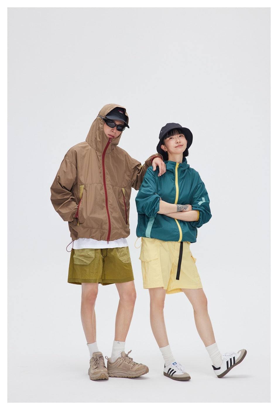 Two models wear hooded jackets: one taupe with red trim and one teal with a yellow zipper.