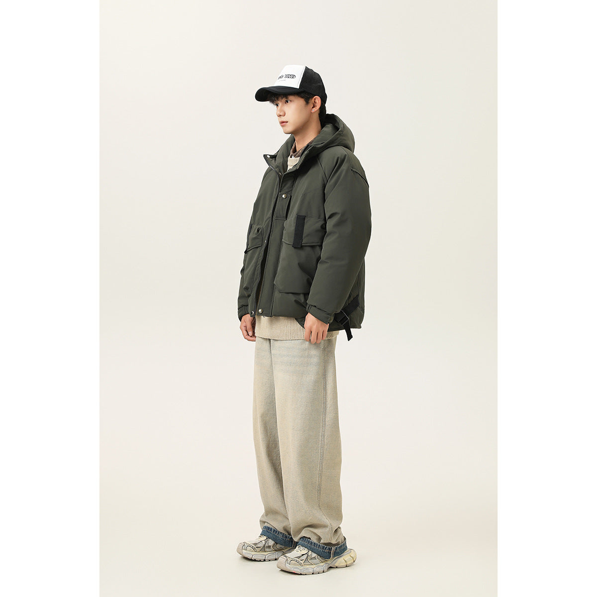 Model in a dark olive hooded jacket with large flap pockets, shown from a side angle with loose light trousers.