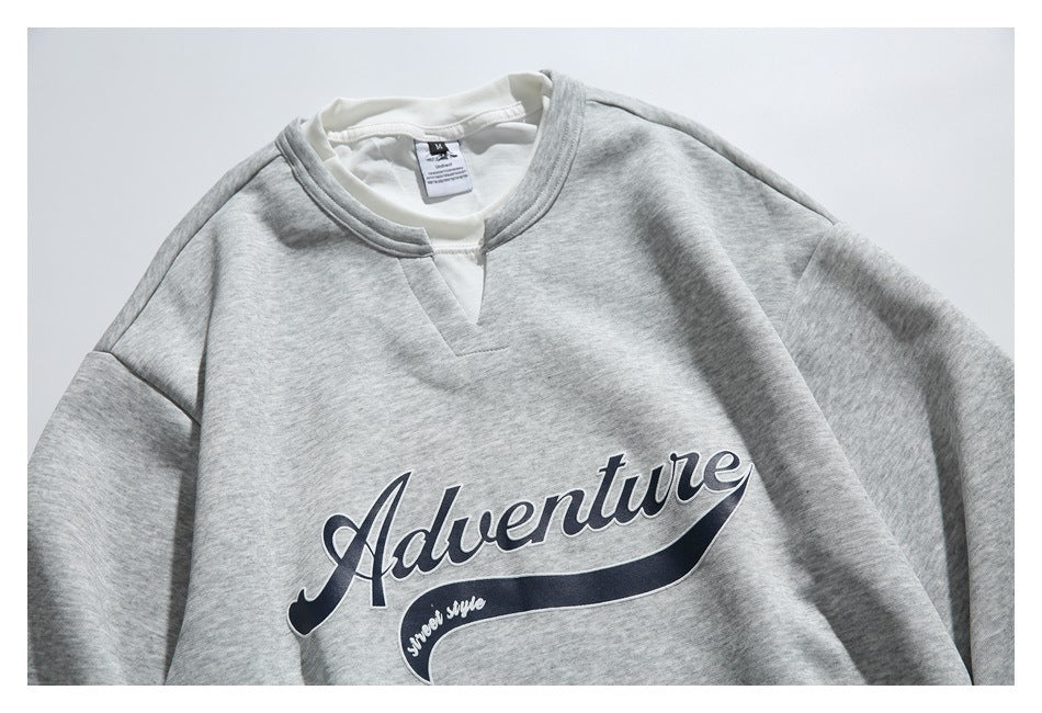 Close view of a light gray sweatshirt front with a split crew neckline and large navy “Adventure” script graphic.