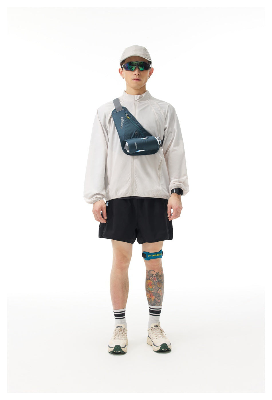 Full-length model wearing a light gray zip jacket and black shorts, with a blue chest bag and cap.