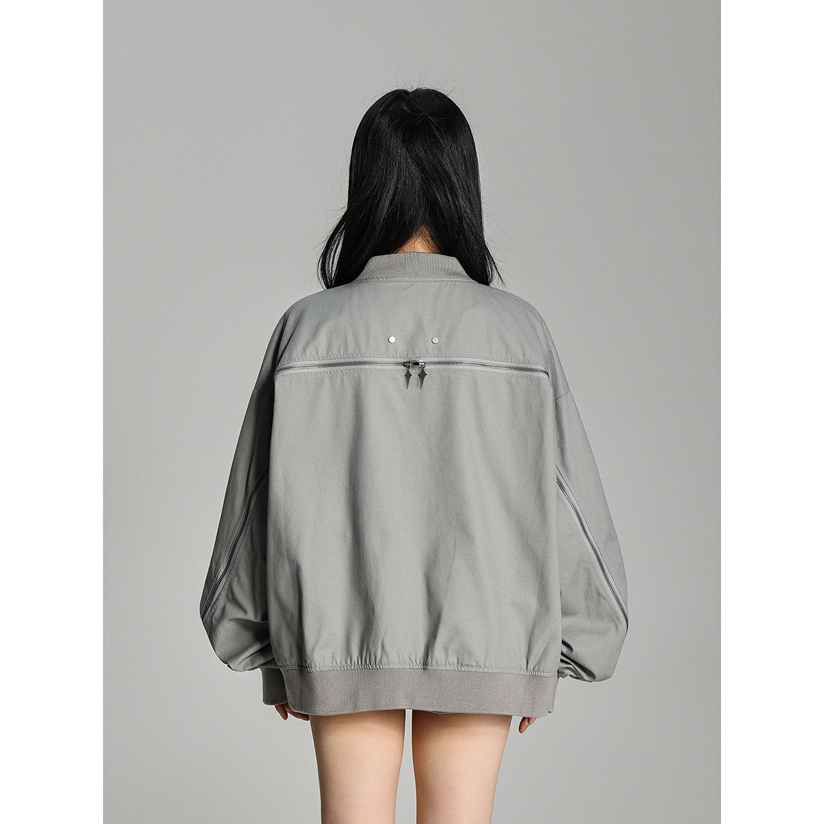 Rear view of a model wearing an oversized gray jacket with a horizontal back zipper and zippered sleeves.