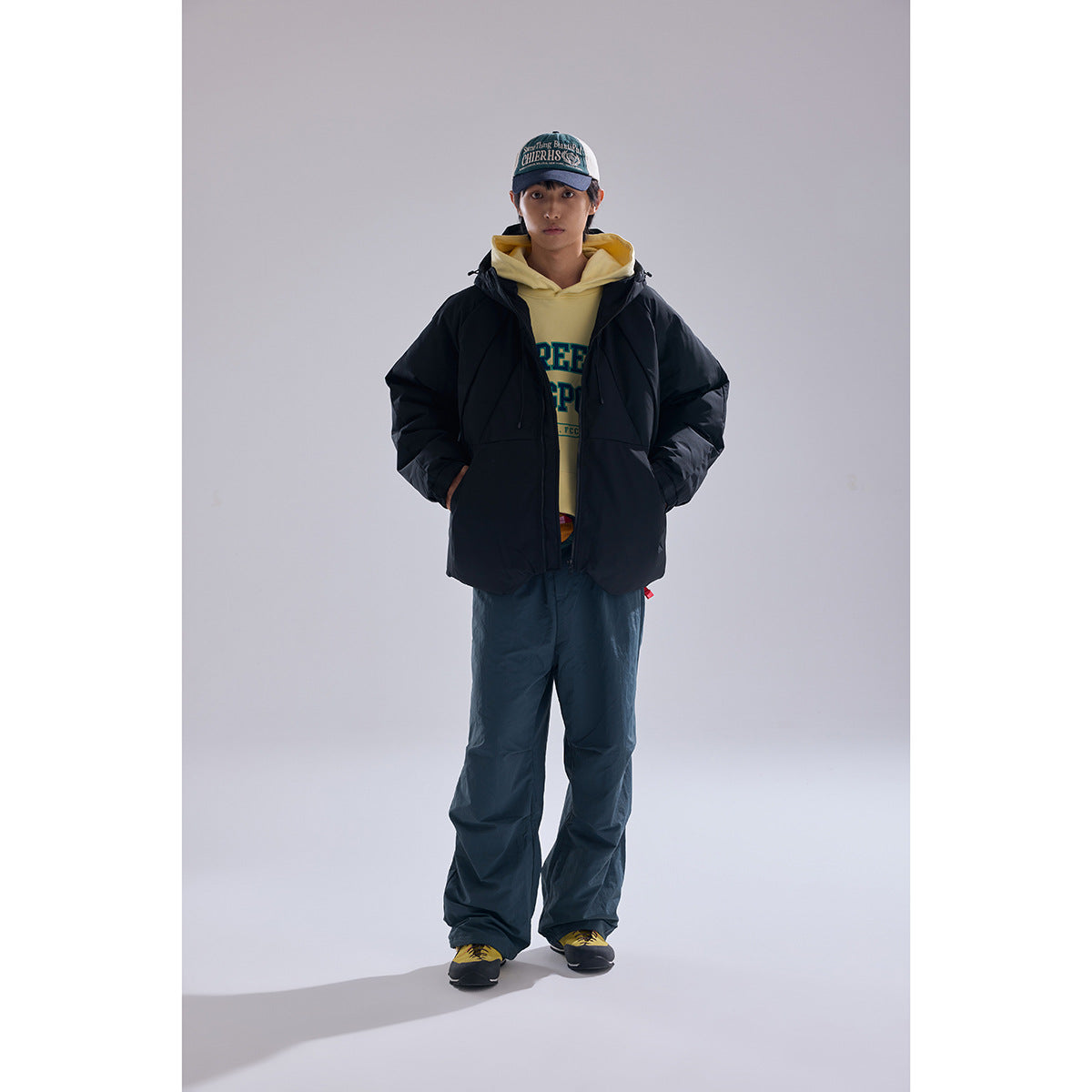 Model wearing a black hooded puffer jacket over a yellow hoodie with dark loose pants, shown from the front.