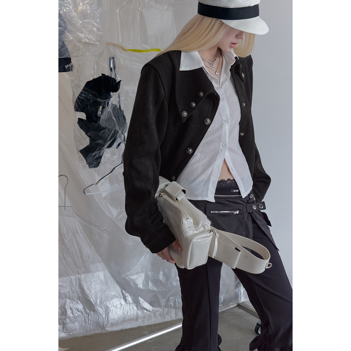 Model wearing a black buttoned jacket with ribbed sleeves over a white shirt, paired with black trousers and a white hat.