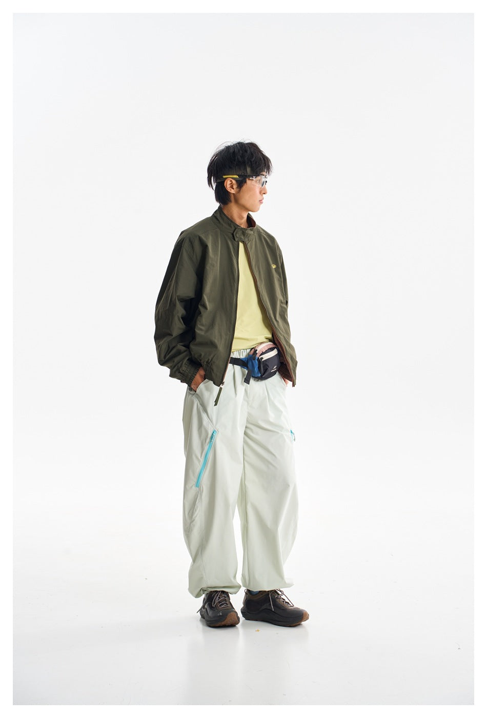 Model wearing a dark olive jacket over a pale yellow shirt and loose white trousers with turquoise zippers.