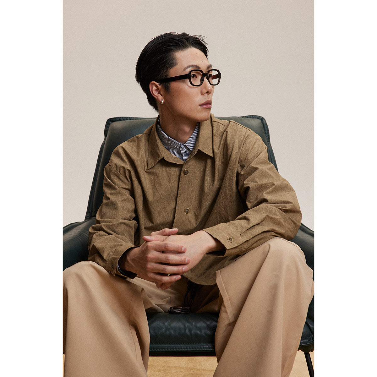 Seated model wearing a brown button-front overshirt over a striped shirt and beige trousers.