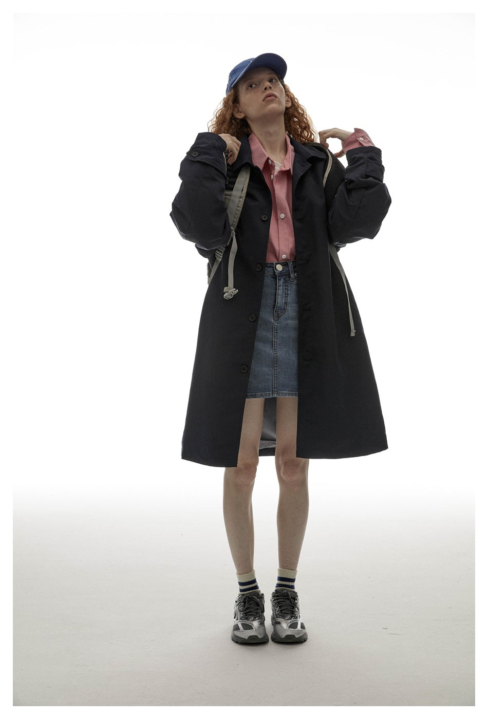 Model wearing a long dark coat over a pink shirt and denim skirt, holding backpack straps.