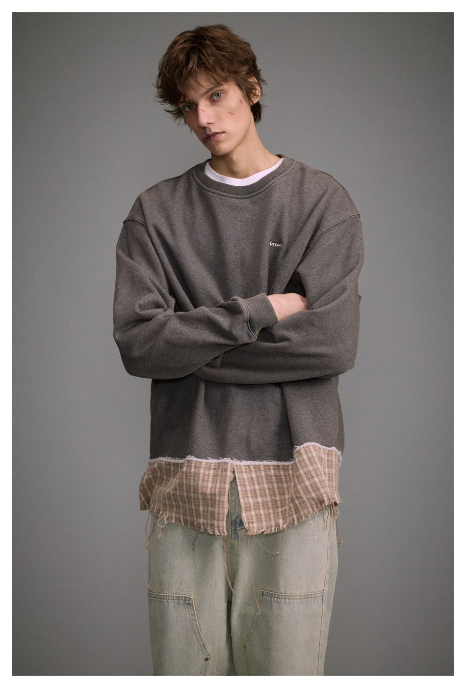 Framed portrait of a man in a charcoal oversized sweatshirt with a frayed plaid hem and loose pale jeans.