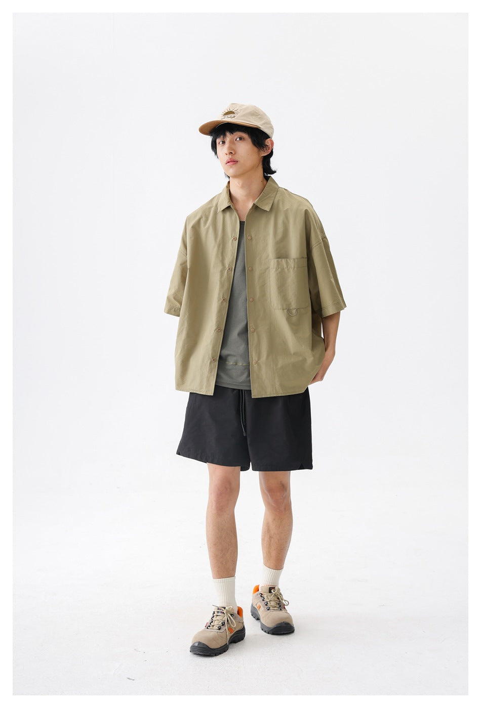 Model wearing an olive-khaki short-sleeve button-up shirt with a chest pocket and black shorts.