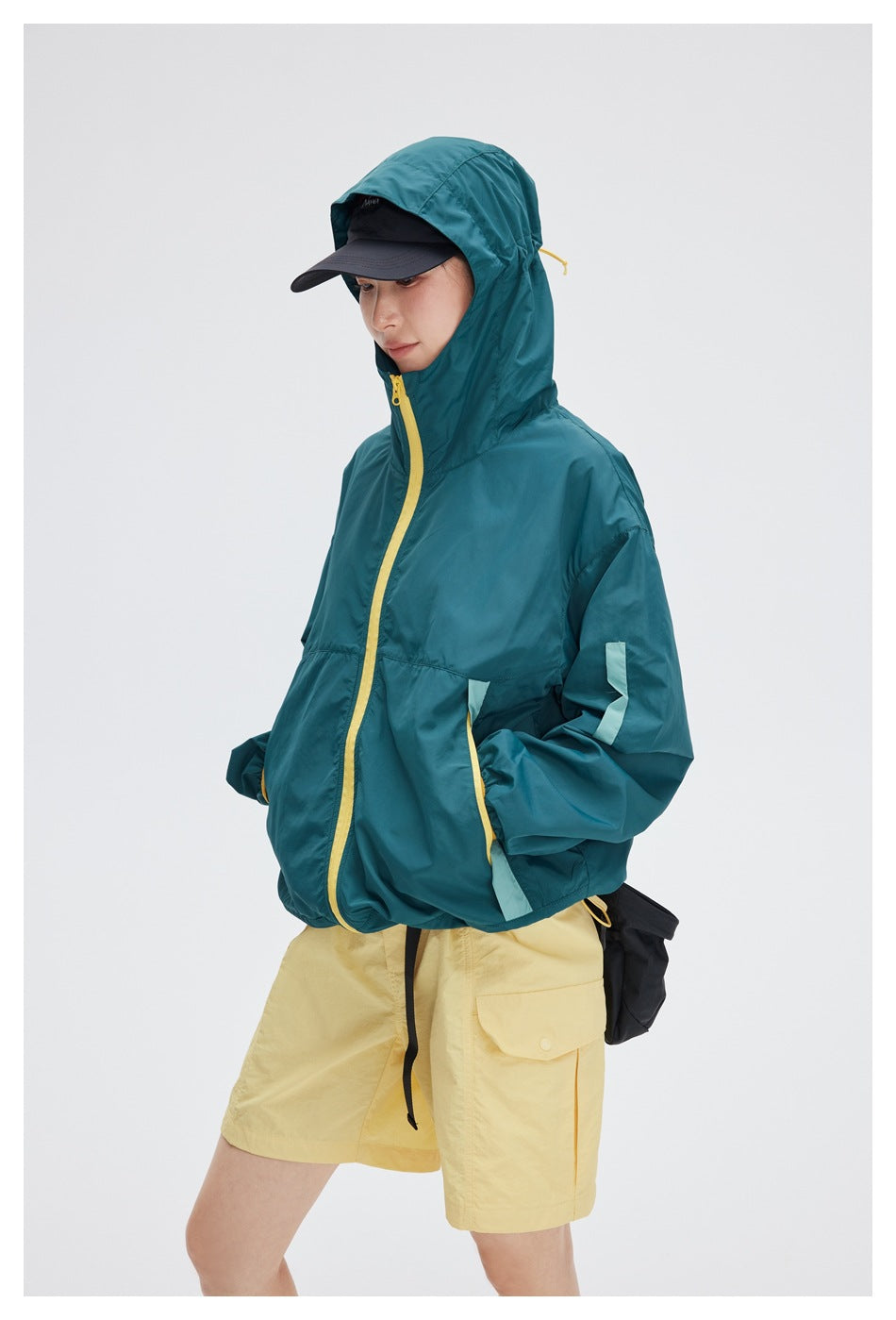 Model wearing a teal hooded jacket with a yellow front zipper and pale yellow shorts.