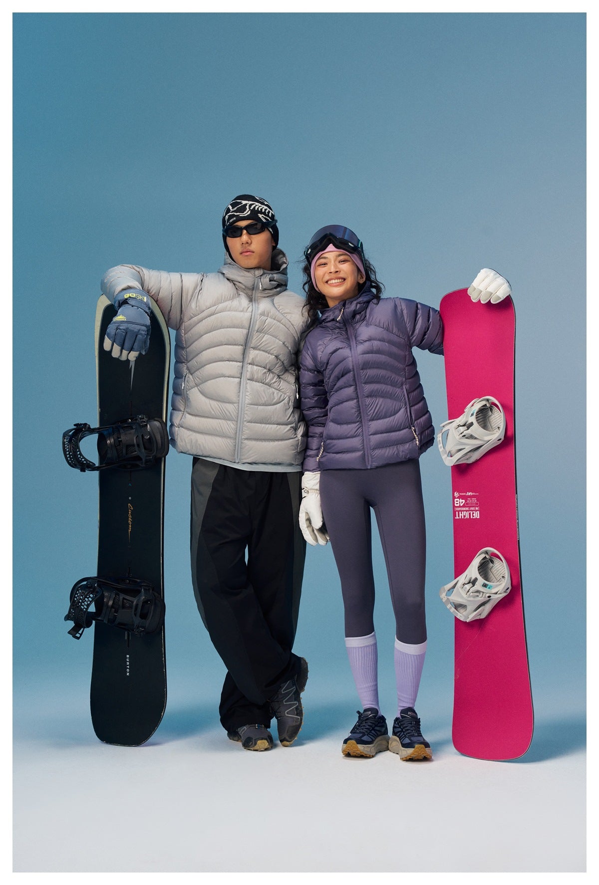 Two snowboarders pose in light gray and lavender quilted puffer jackets beside their snowboards.