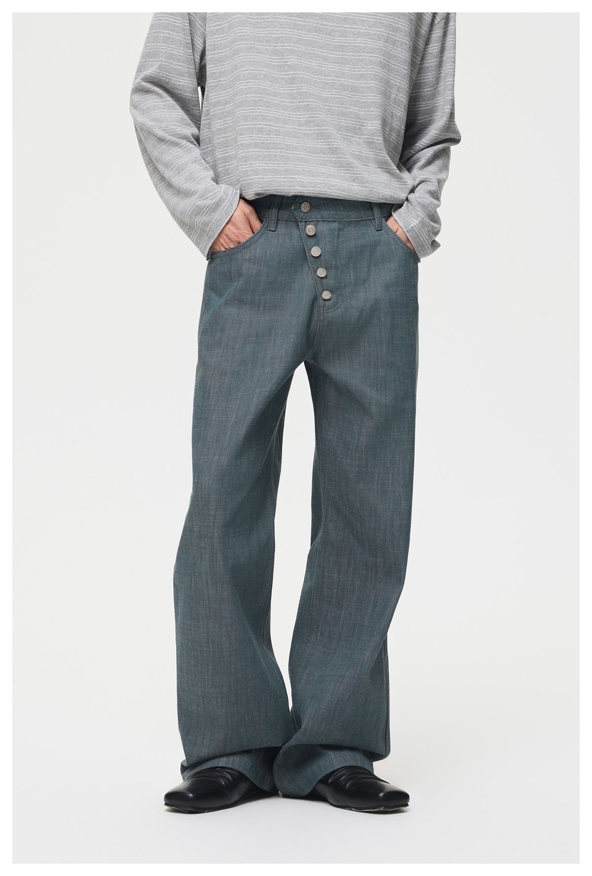 Model wearing blue-teal wide-leg jeans with diagonal button front, gray striped top and black shoes, hands in pockets