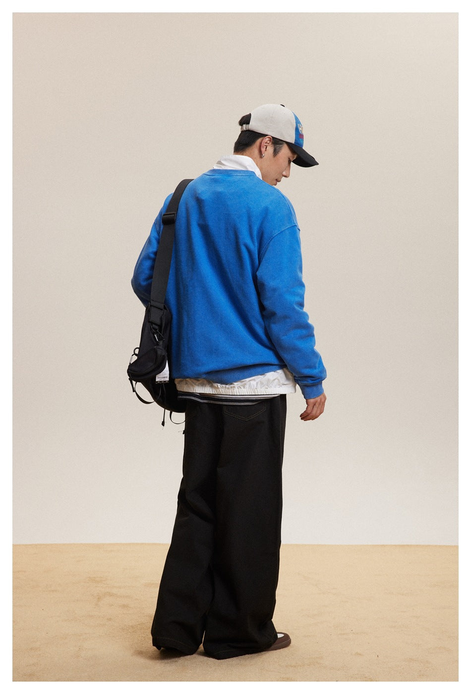 Back view of a model wearing a bright blue sweatshirt and wide black trousers with a shoulder bag.